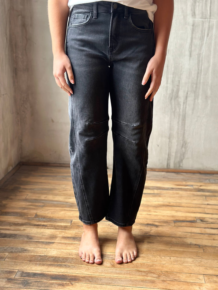 A person wearing dark washed denim jeans with a straight-leg or slightly relaxed fit, standing barefoot on a wooden floor against a textured concrete wall.