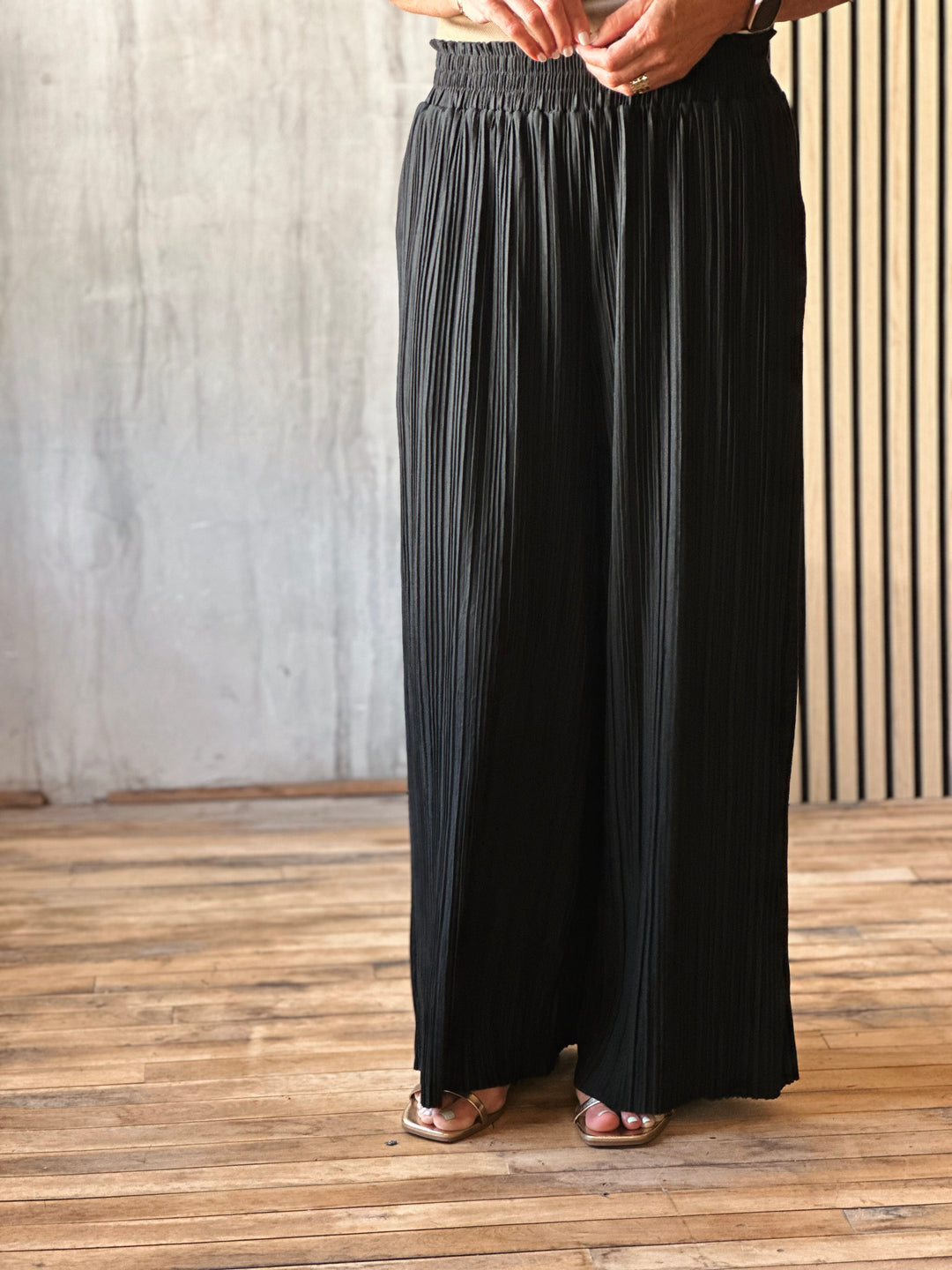 Person wearing black wide-leg pants standing on a wooden floor with a light gray wall background
