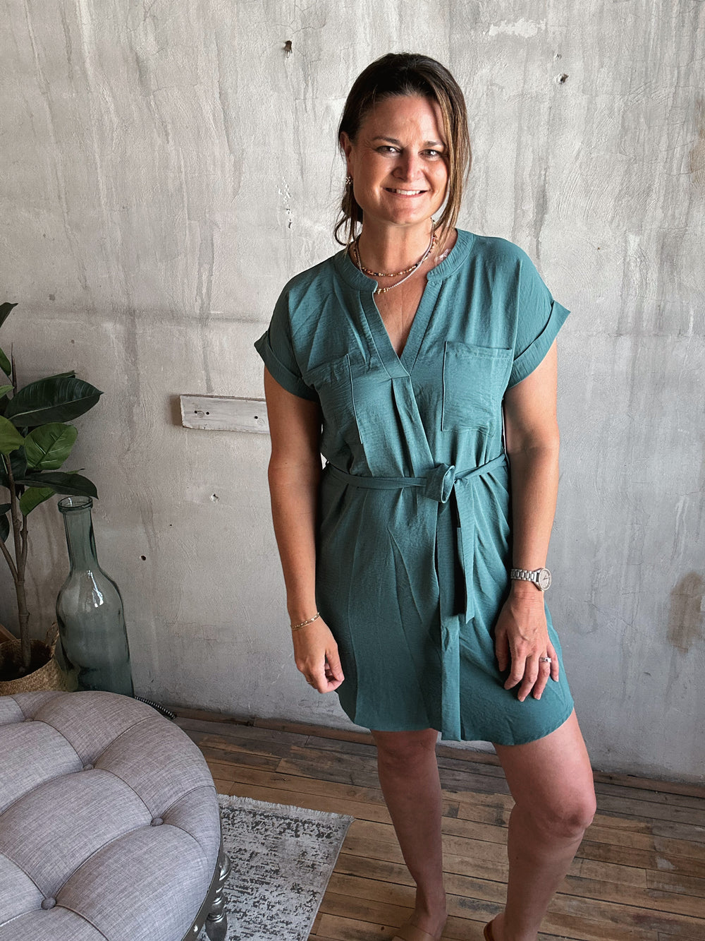 A woman wearing a dark teal dress with strings to tie in the front and pockets standing in a room with a gray wall and a plant.