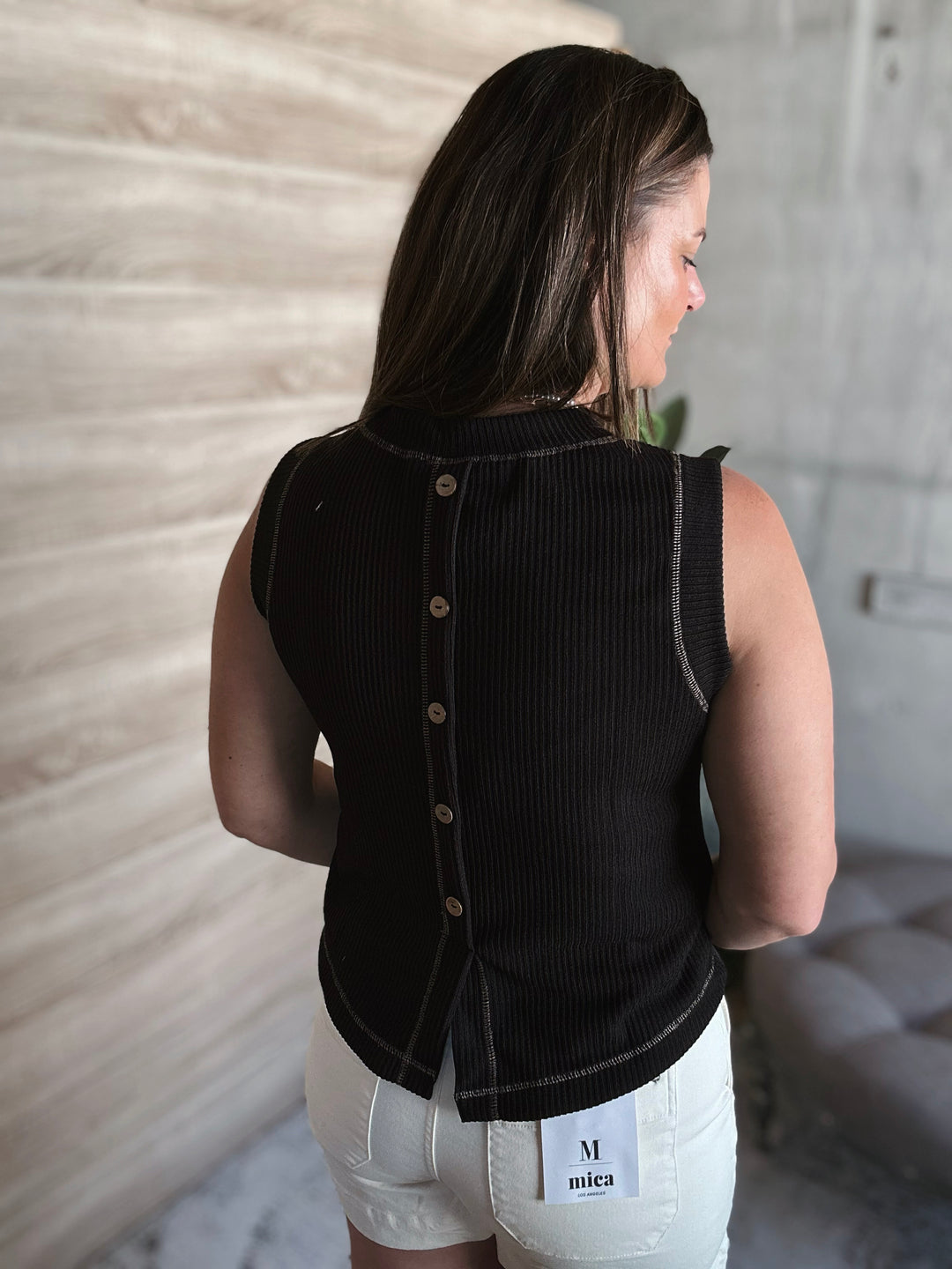 Woman wearing a black sleeveless top with buttons going down the middle of the back on the shirt with a blurred background.