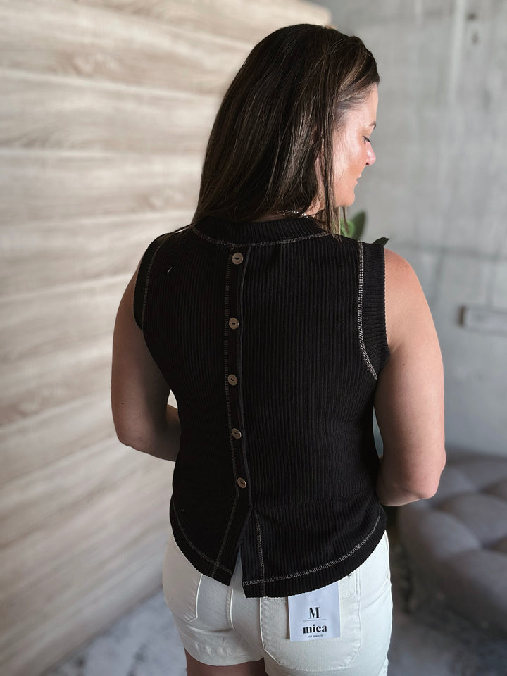 Woman wearing a black sleeveless top with buttons going down the middle of the back on the shirt with a blurred background.