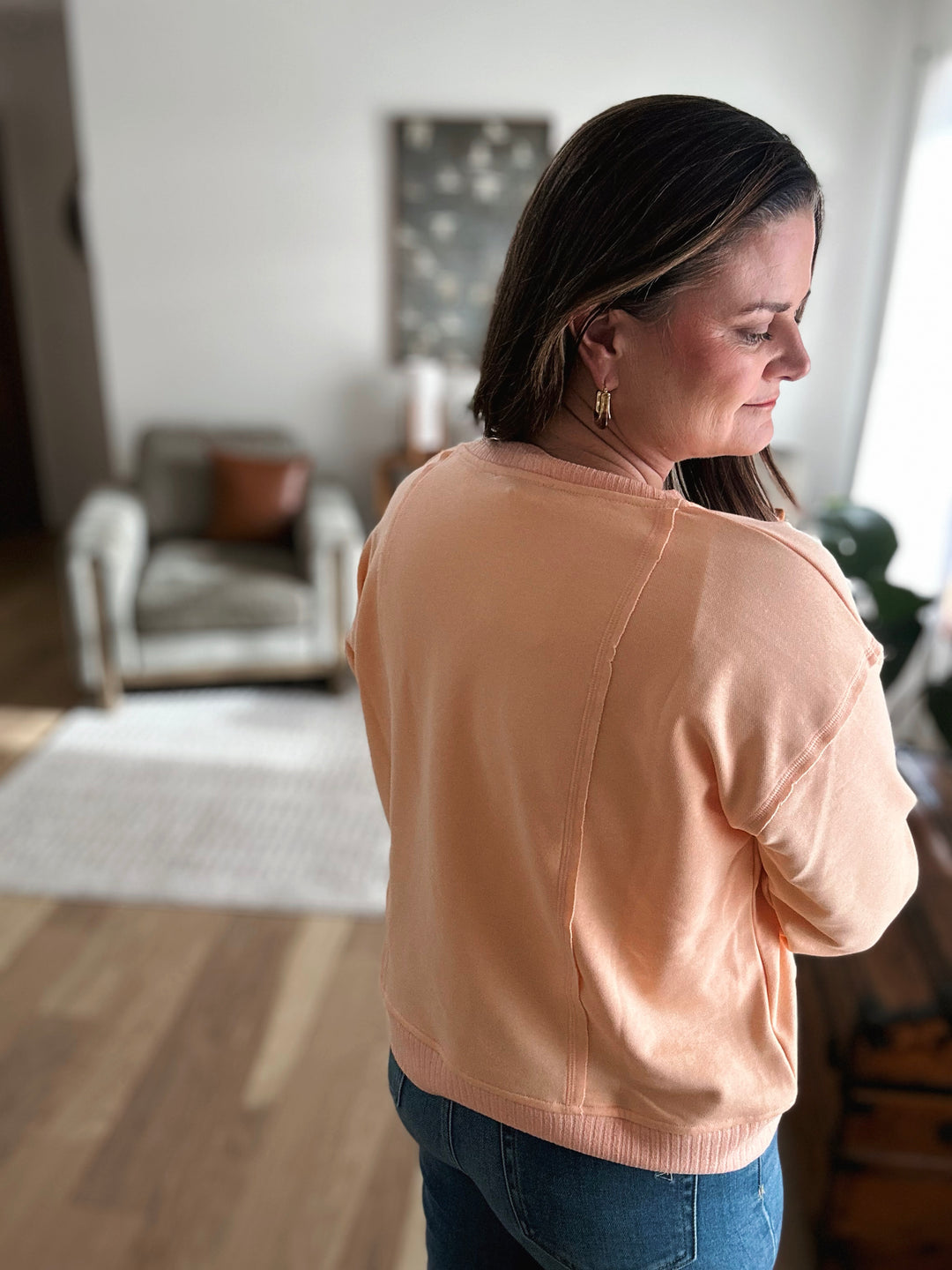 Woman wearing a peach-colored sweater in a room with a chair and rug.