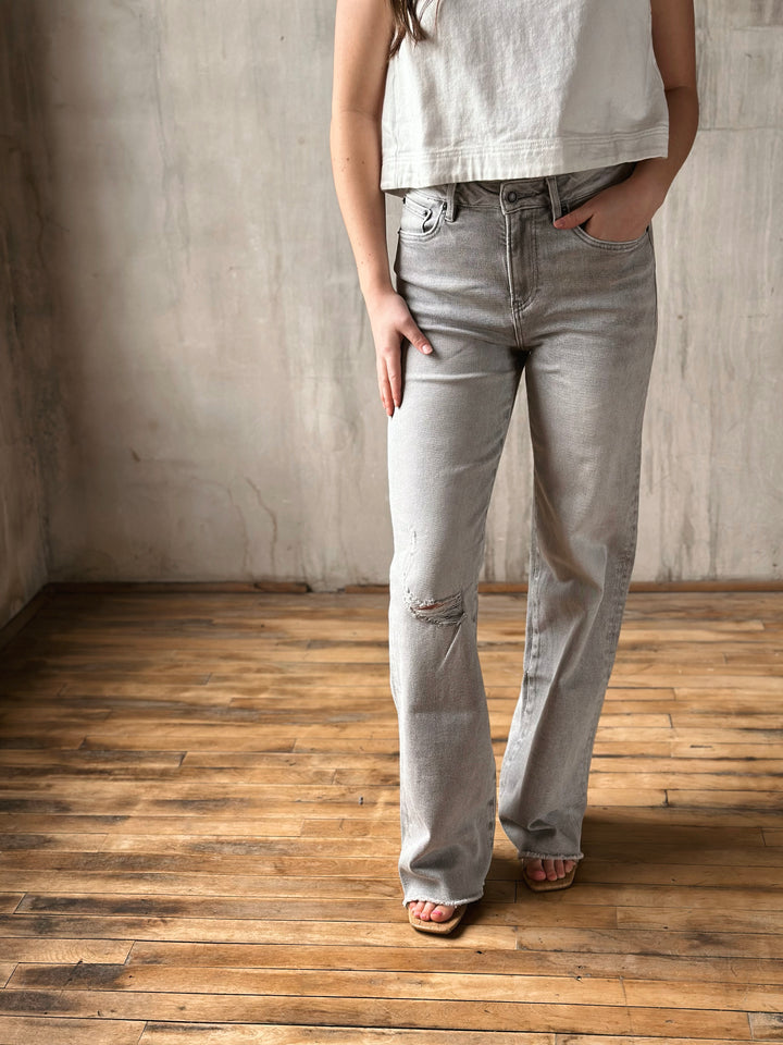 Person wearing light gray jeans and a white top standing on a wooden floor with a concrete wall background.