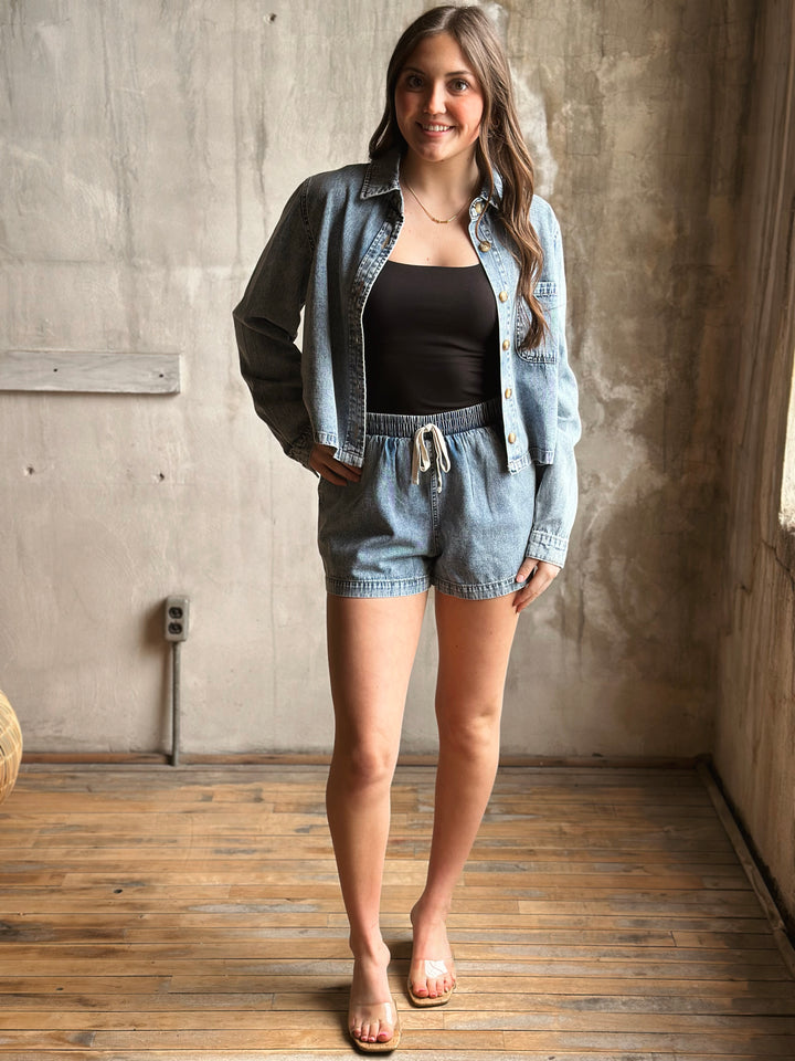 Woman wearing a denim jacket and shorts standing in a room with wooden floor and concrete wall.