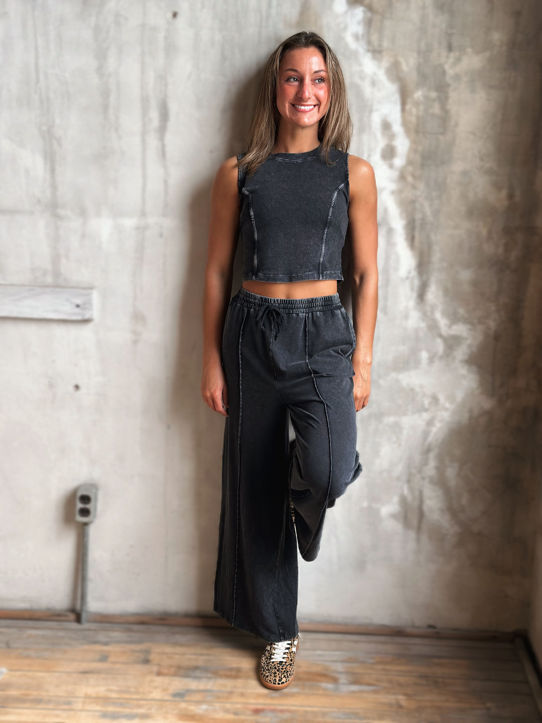 Woman wearing a black sleeveless top and wide-leg pants standing against a textured wall.