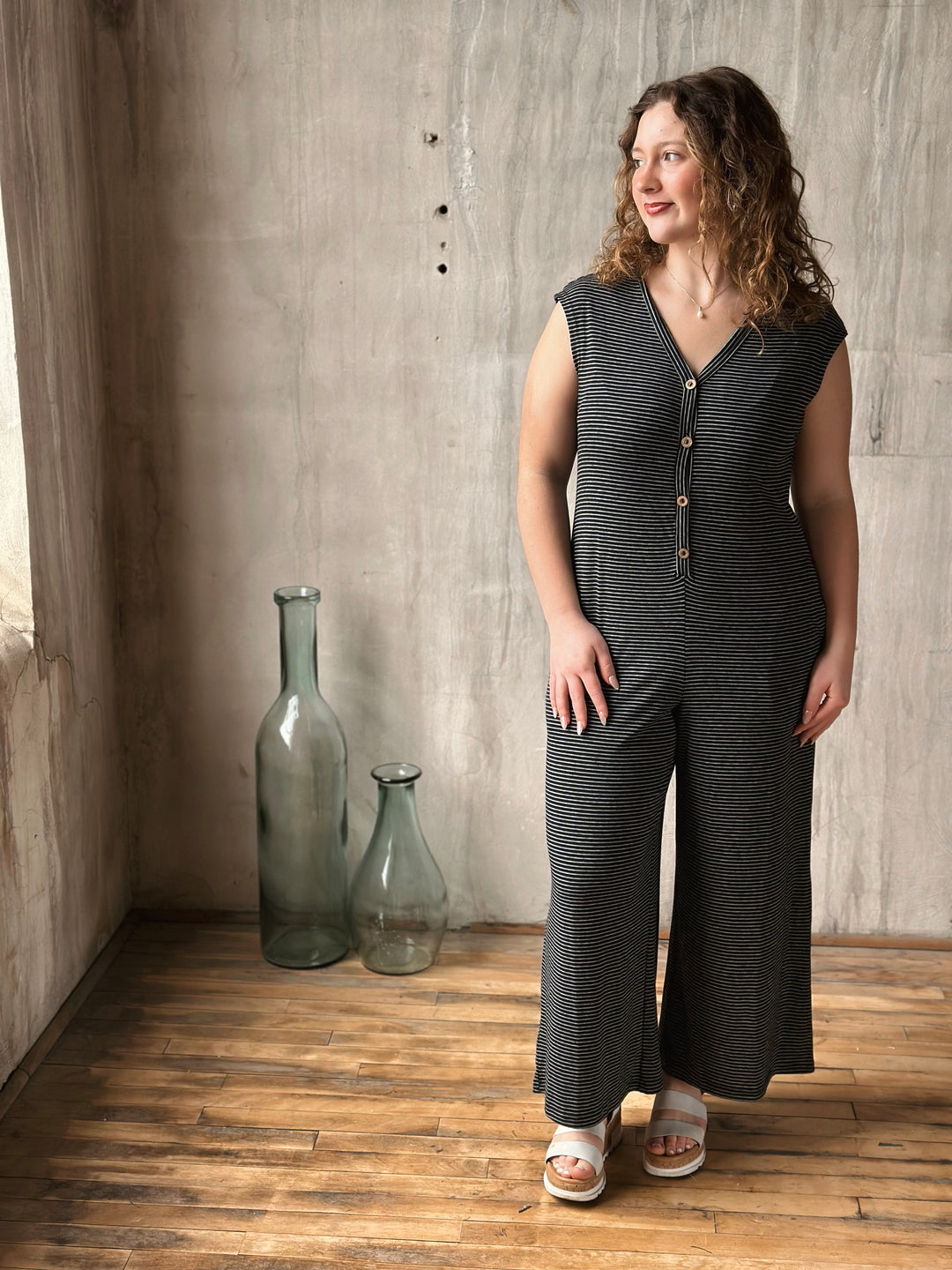 Woman wearing a black striped sleeveless jumpsuit standing in a room with wooden floor and concrete wall.