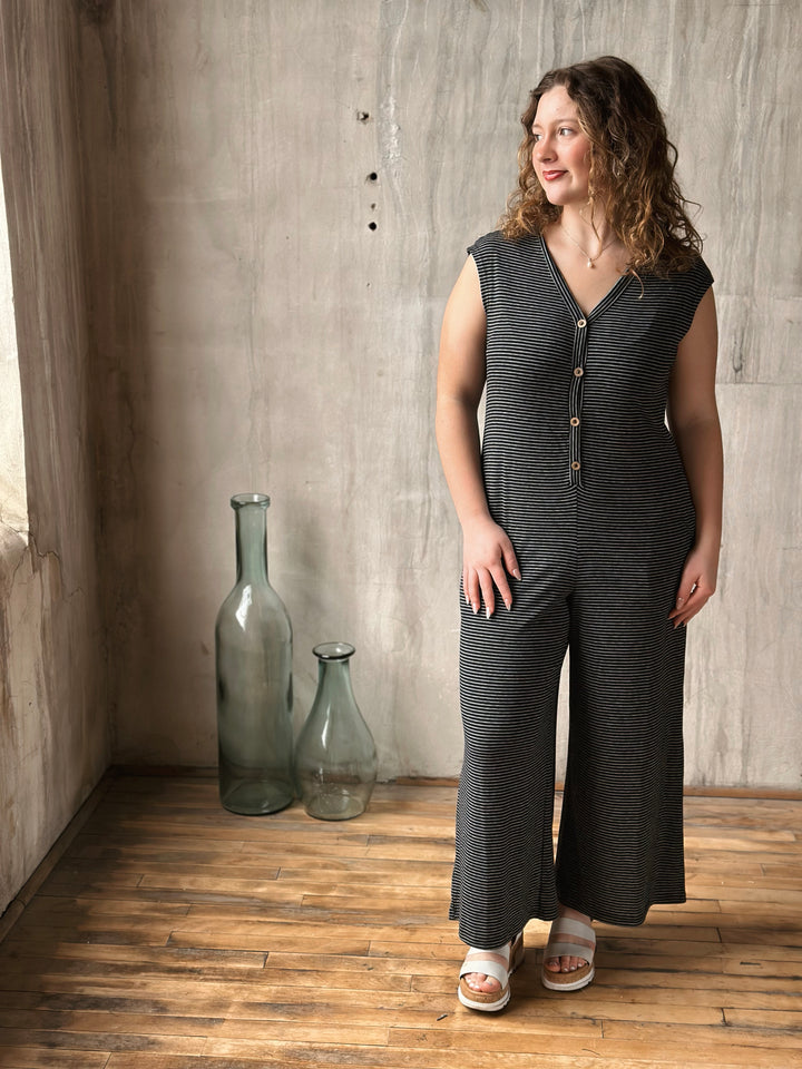 Woman wearing a black striped sleeveless jumpsuit standing in a room with wooden floor and concrete wall.
