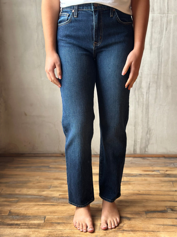 A person wearing dark blue straight-leg denim jeans, standing barefoot on a wooden floor against a textured concrete wall.