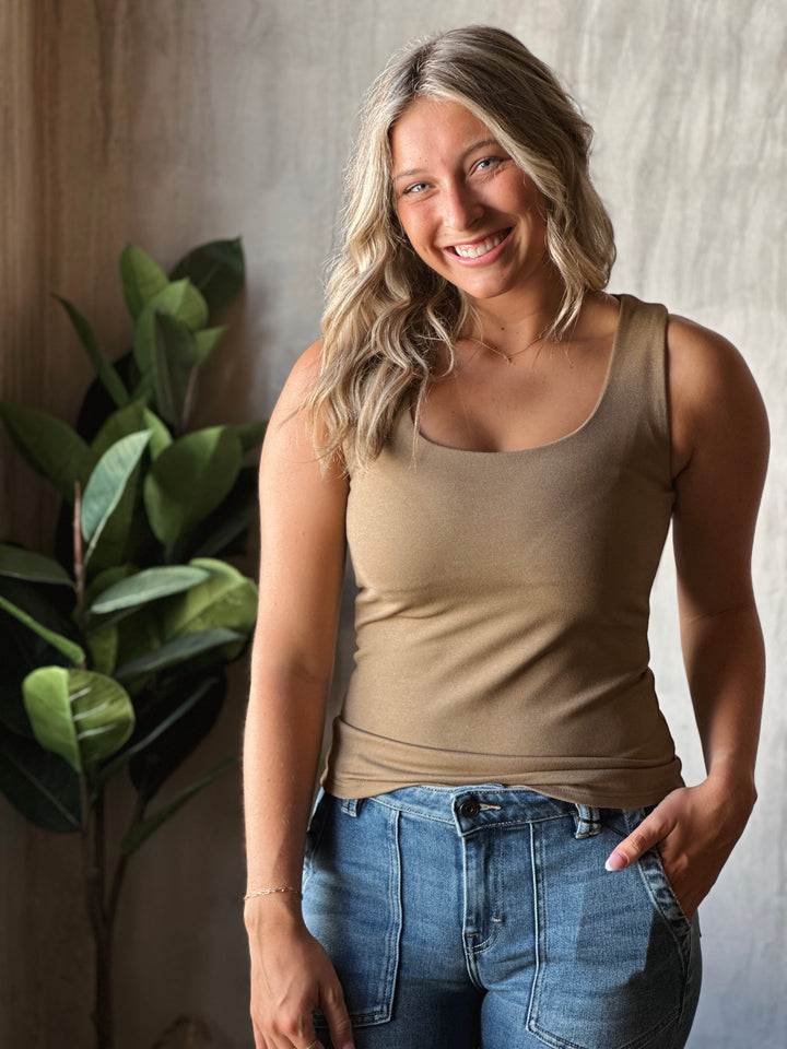 Woman wearing a beige tank top and blue jeans standing against a neutral wall with a plant to the left.