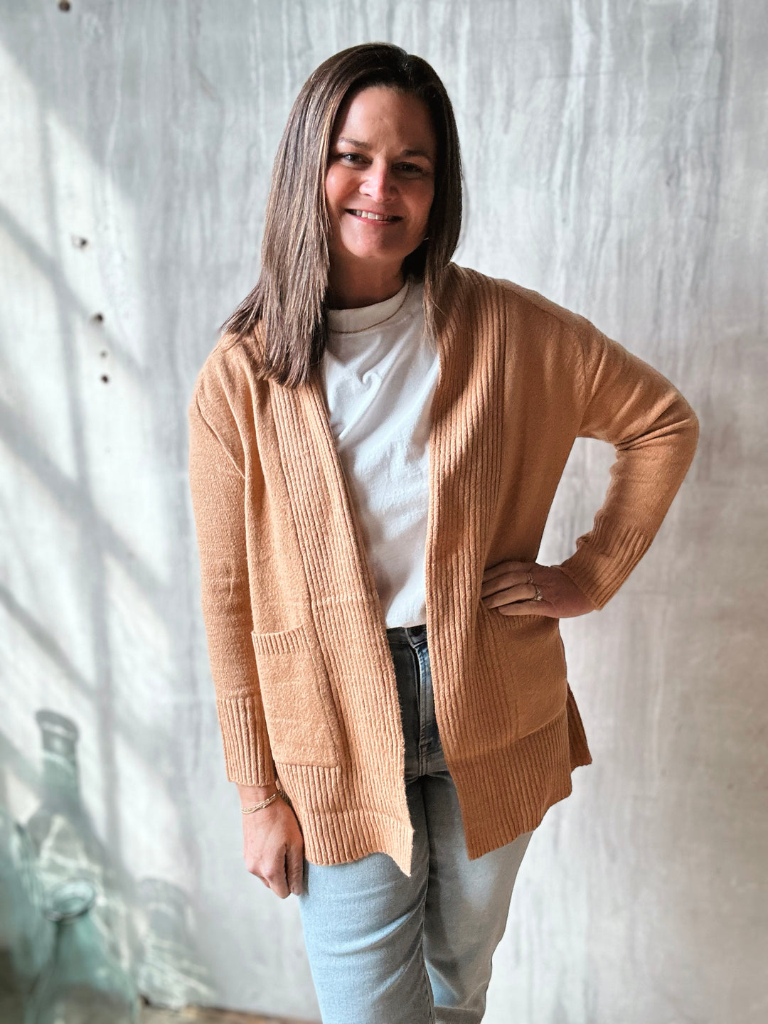 Woman wearing a beige cardigan over a white shirt and light blue jeans, standing against a textured white wall.