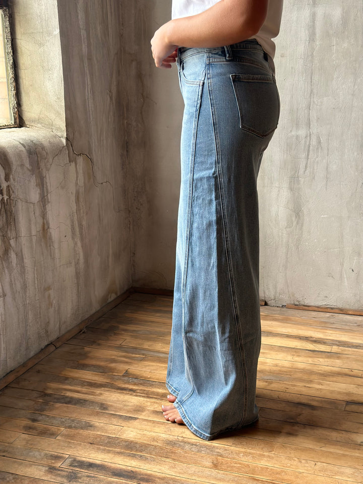 A person wearing light-wash blue denim jeans with a wide-leg, flared silhouette, standing barefoot on a wooden floor against a textured concrete wall.