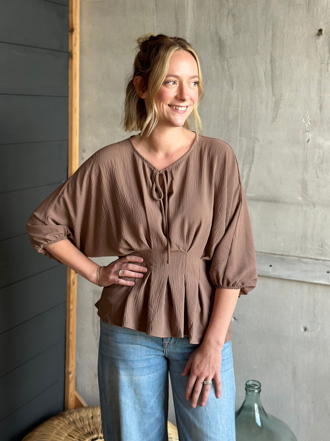 Woman wearing a brown blouse and blue jeans standing against a concrete wall.