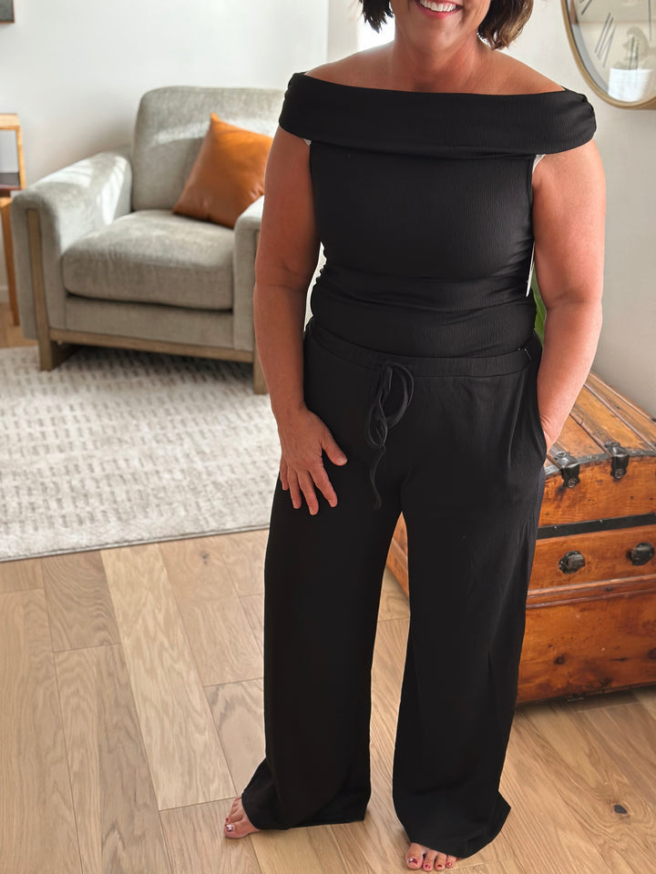 Woman wearing a black off-shoulder top and wide-leg pants in a living room.