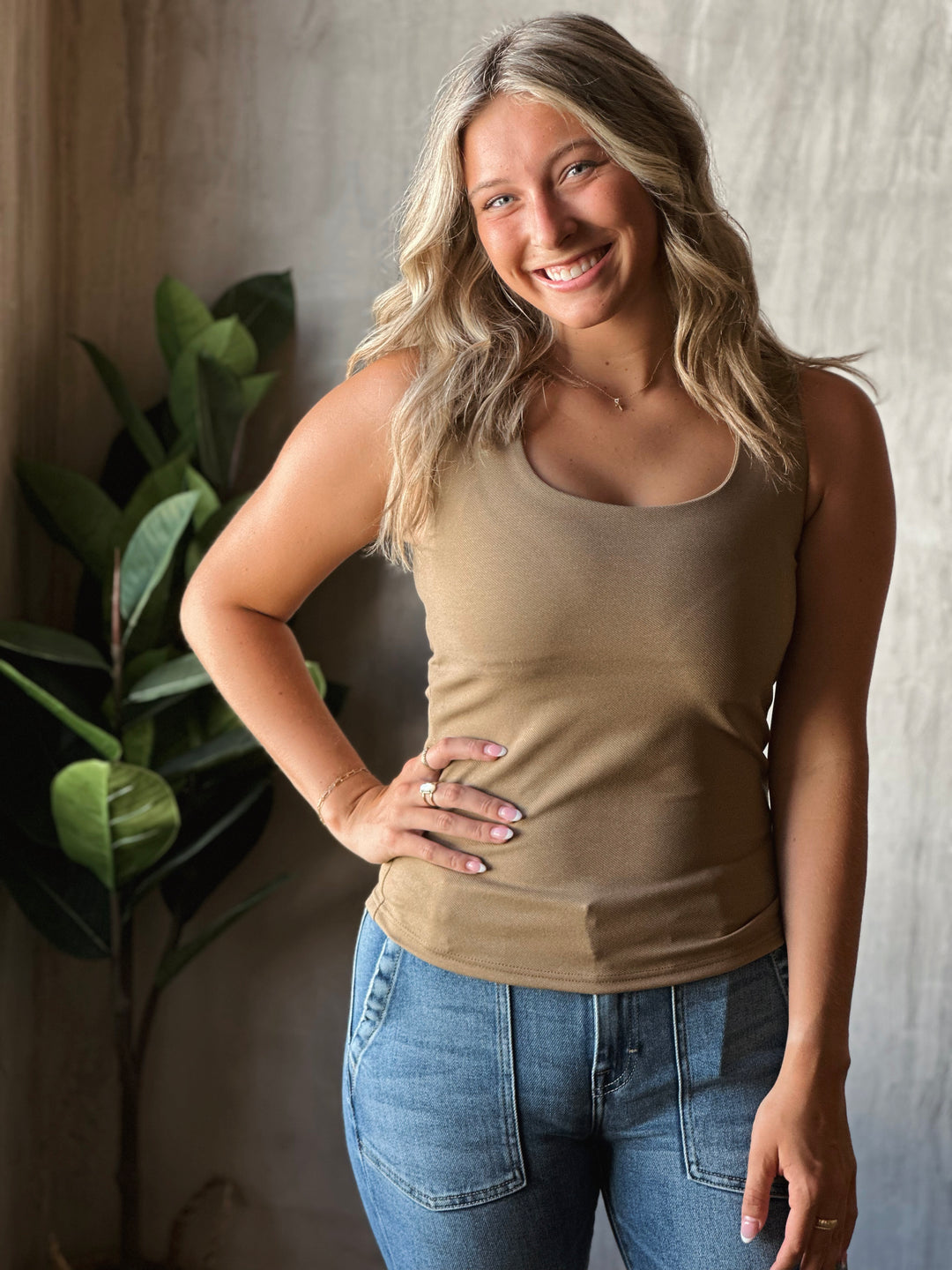Woman wearing a beige tank top and blue jeans standing against a neutral background with a plant.