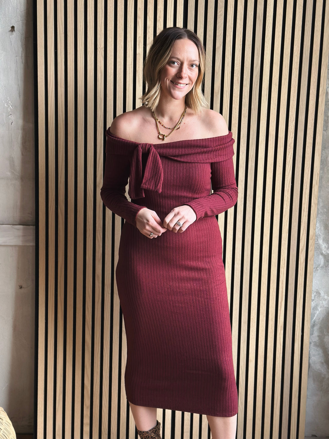 Woman wearing a burgundy off-shoulder dress standing against a striped wall.