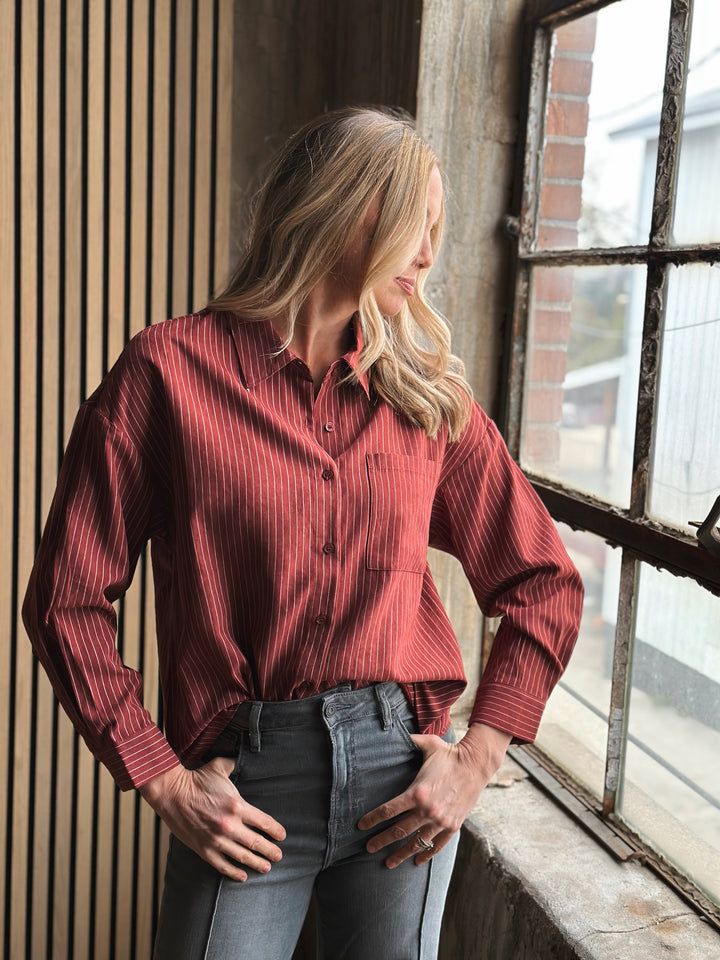 Woman wearing a red shirt and gray pants leaning against a window.