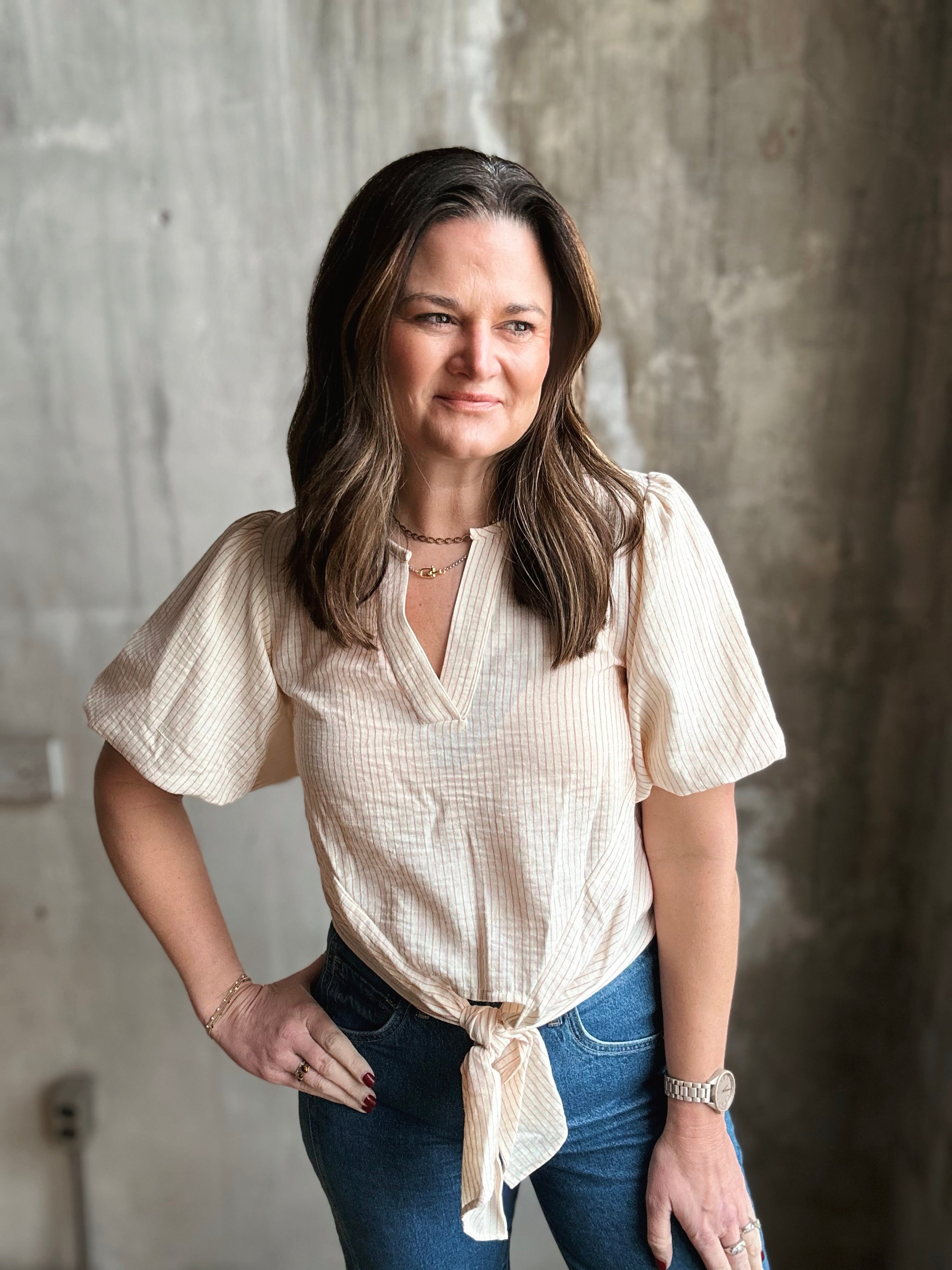 Woman wearing a beige blouse and blue jeans against a textured wall.