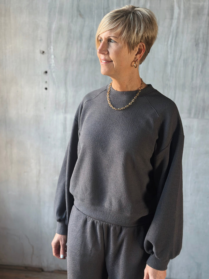 Woman wearing a dark gray sweater and pants against a textured wall.