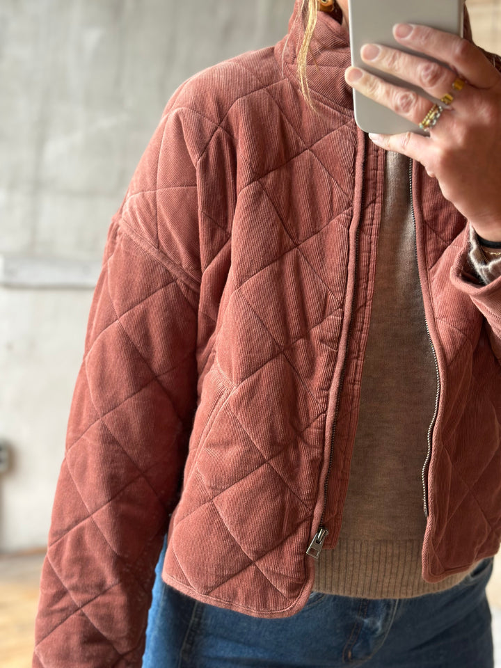 A person wearing a dusty rose quilted jacket over a beige sweater and blue jeans, taking a mirror selfie with a phone visible in the frame.