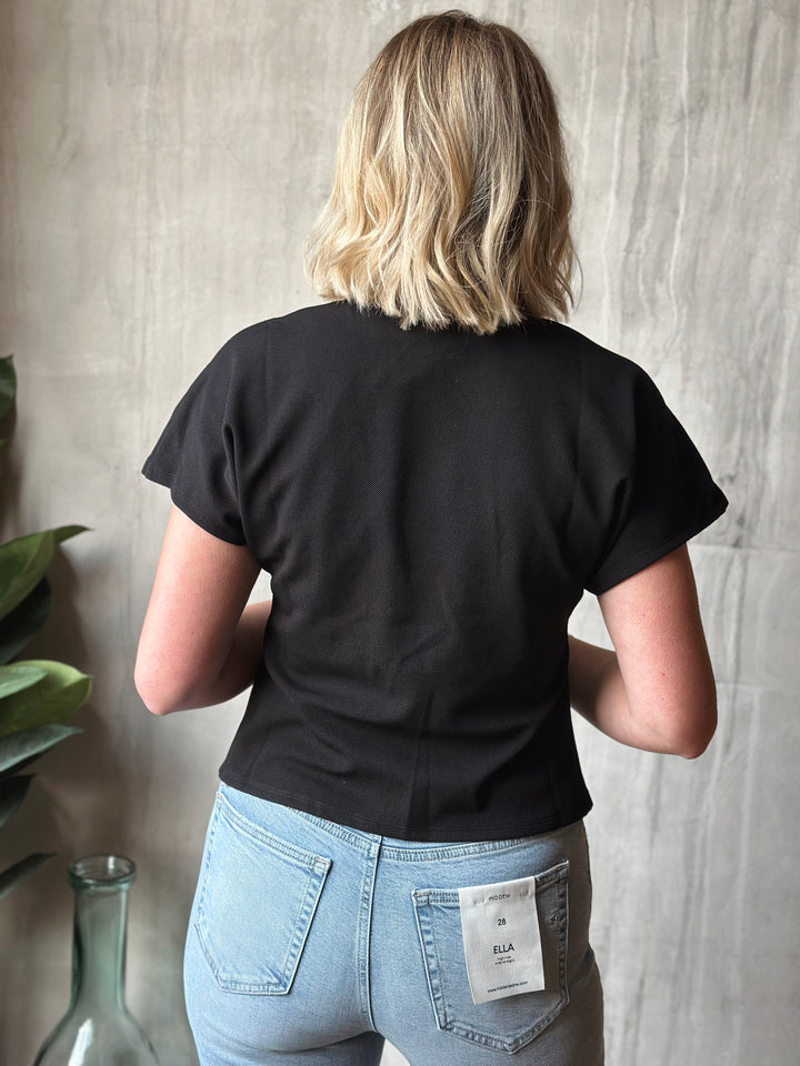 Person wearing a black t-shirt and light blue jeans with a visible brand label, standing against a neutral wall.