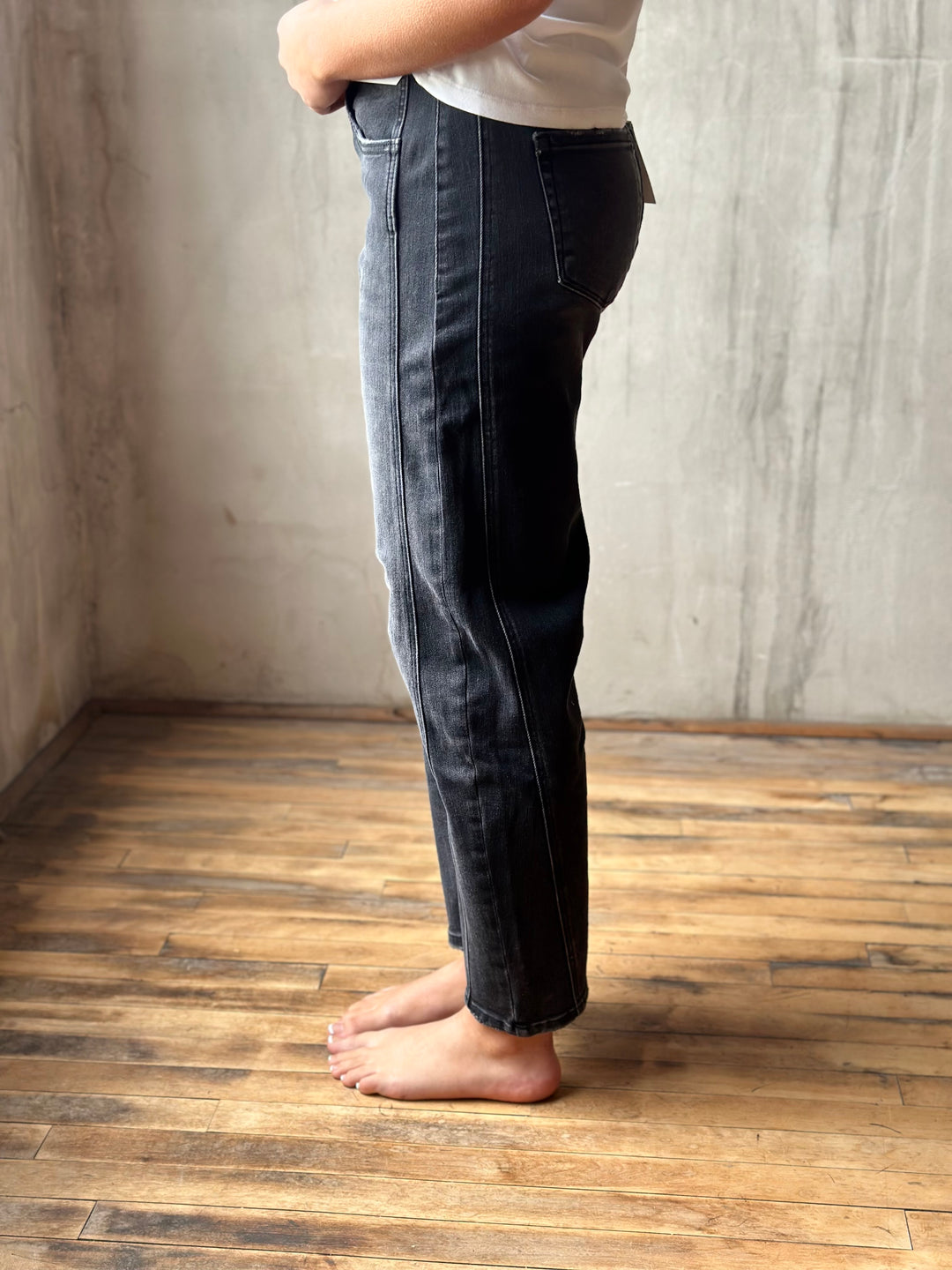 A person shown from the waist down wearing dark indigo denim jeans, standing barefoot on a wooden floor against a textured concrete wall.