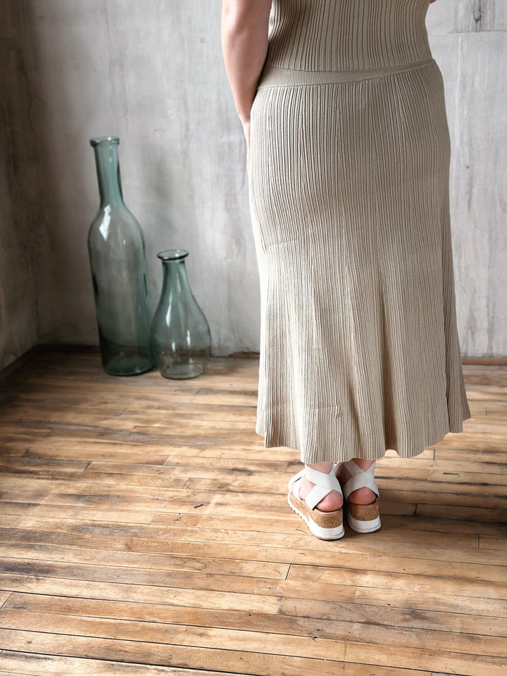 Person wearing a beige ribbed skirt standing on a wooden floor with two glass bottles in the background.