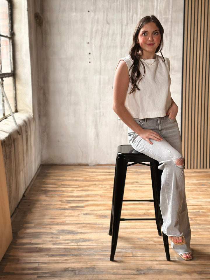 Woman wearing a light gray jeans and a white top in a room with wooden floor and textured walls.