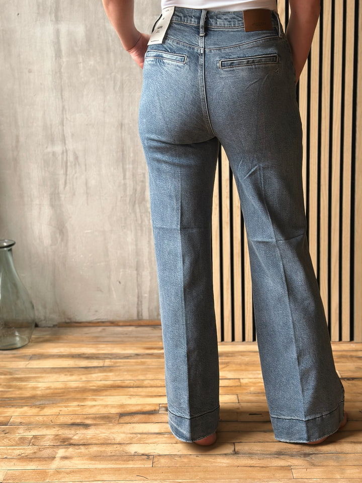 Person wearing blue jeans standing against a wooden floor and wall.
