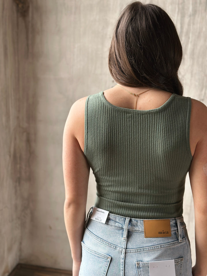 Person wearing a green ribbed bodysuit and light blue jeans against a neutral wall.