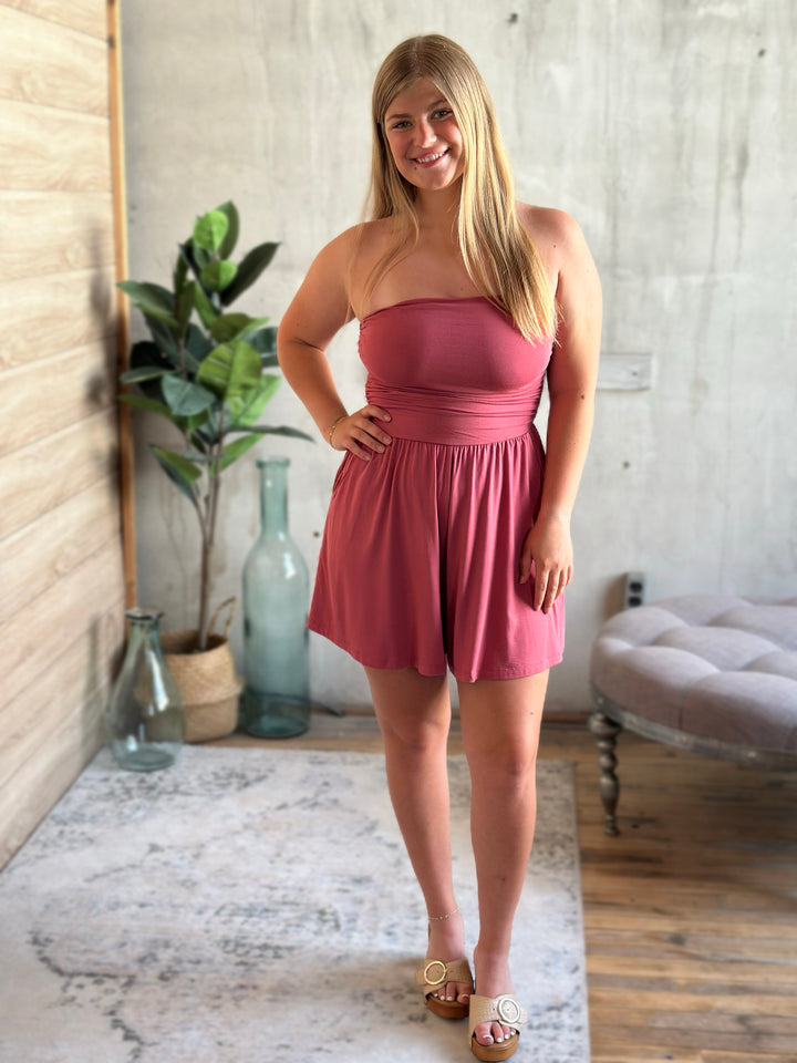 Woman wearing a dark pink strapless romper standing in a room with a plant and a bench.
