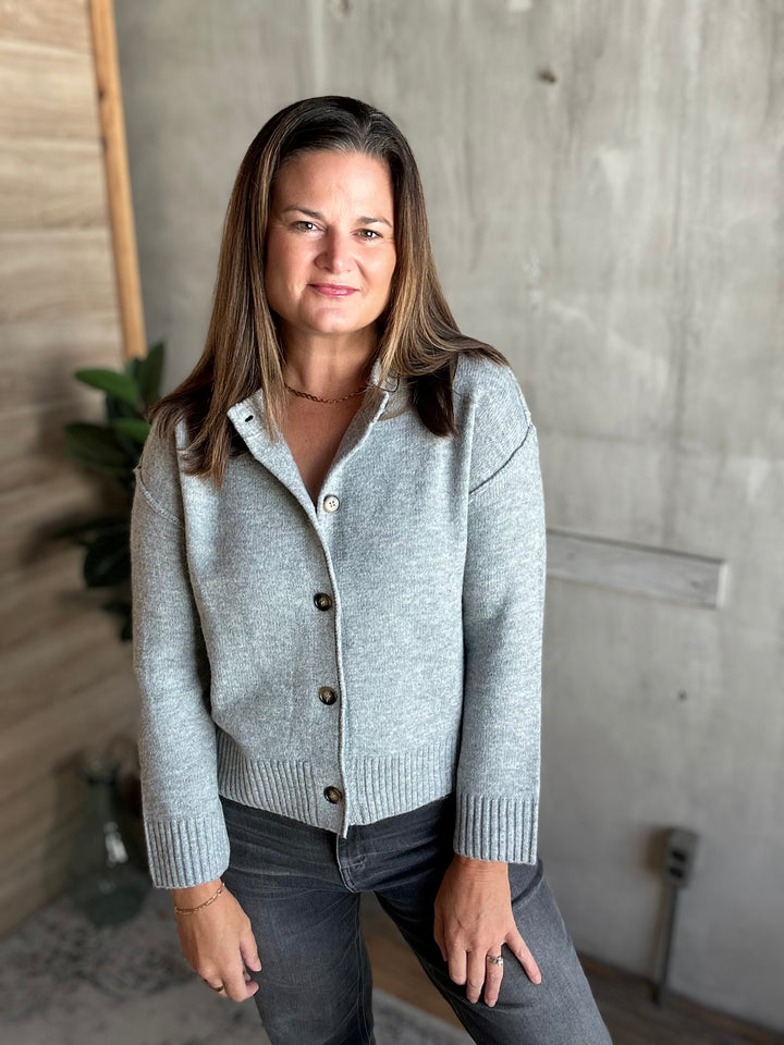 A woman wearing a light gray knit button-front cardigan sweater and dark gray jeans, standing in an indoor setting with a concrete wall and wood paneling in the background.