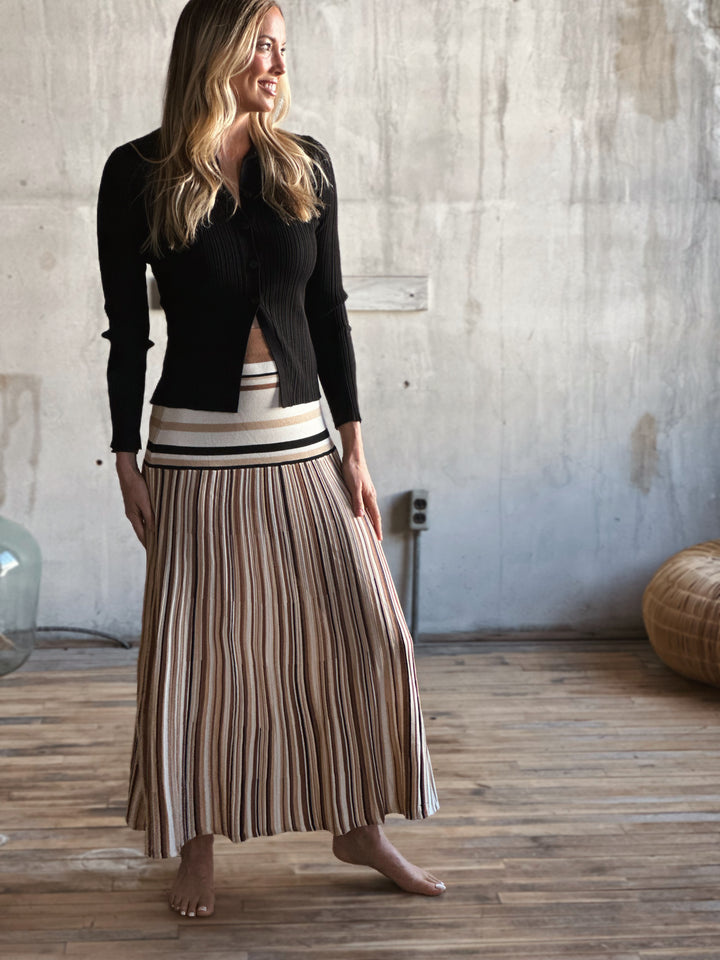 Woman wearing a black top and striped skirt standing in a room with wooden floor and textured wall.