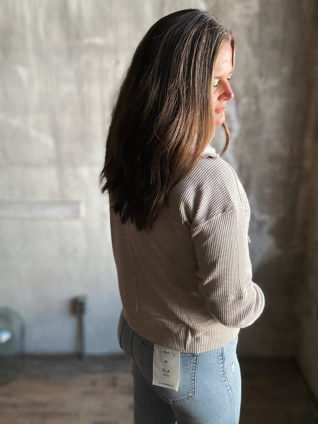 Woman wearing a beige ribbed sweater and light blue jeans with a visible tag, standing against a textured wall.