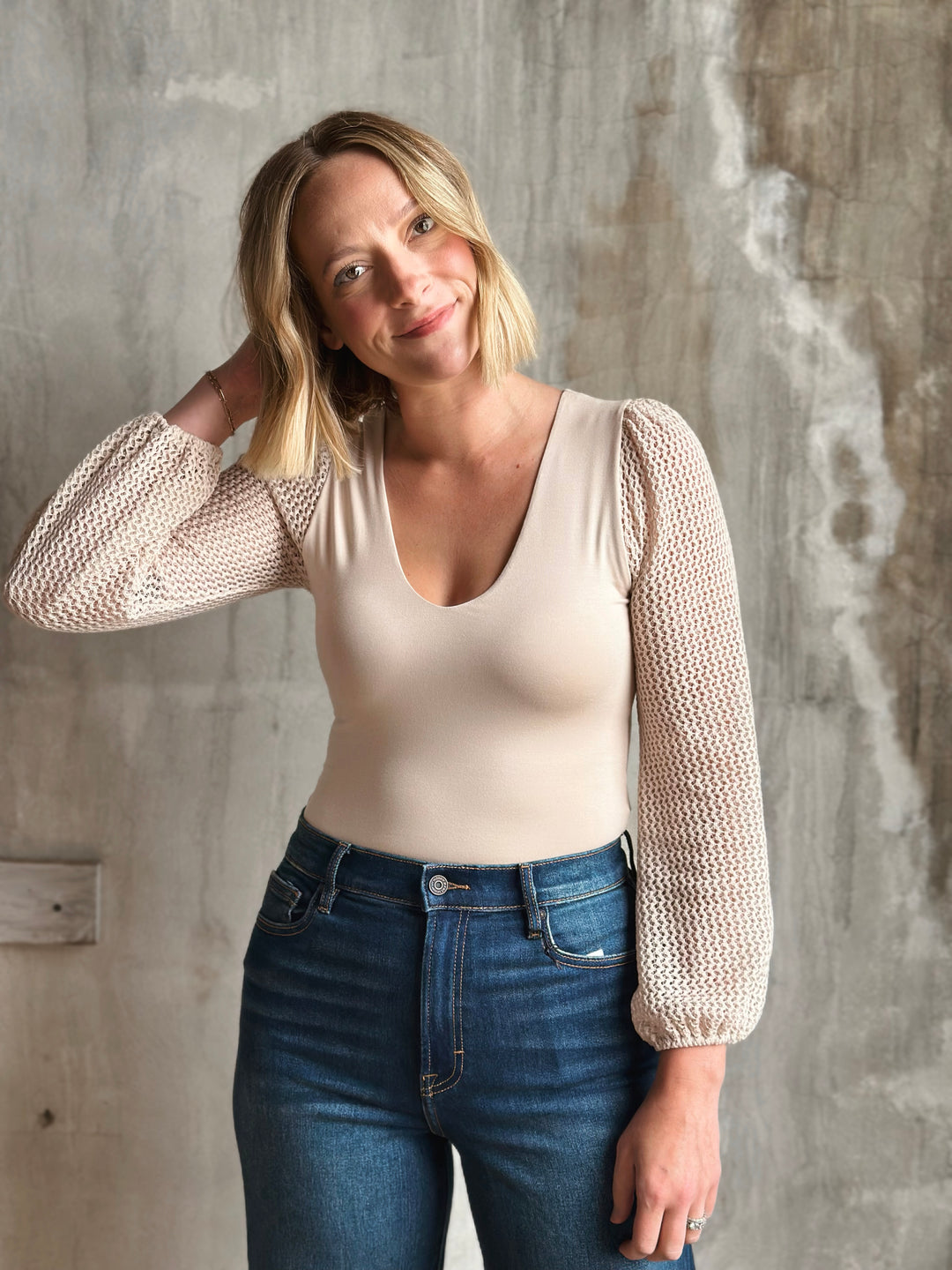 Woman wearing a beige knit cardigan over a white top and blue jeans against a textured wall.