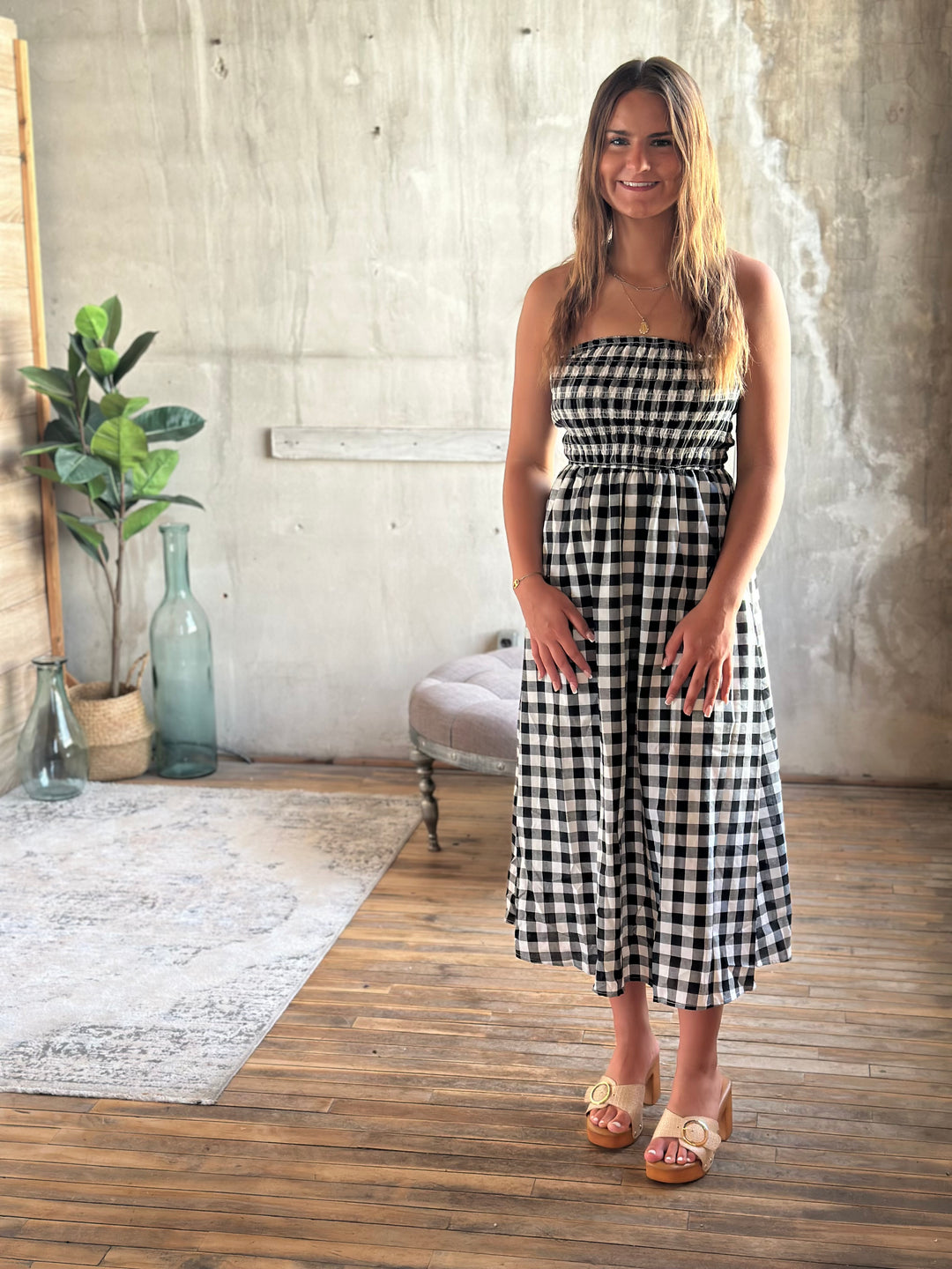 Woman wearing a black and white plaid strapless dress standing in a room with wooden flooring and a concrete wall.