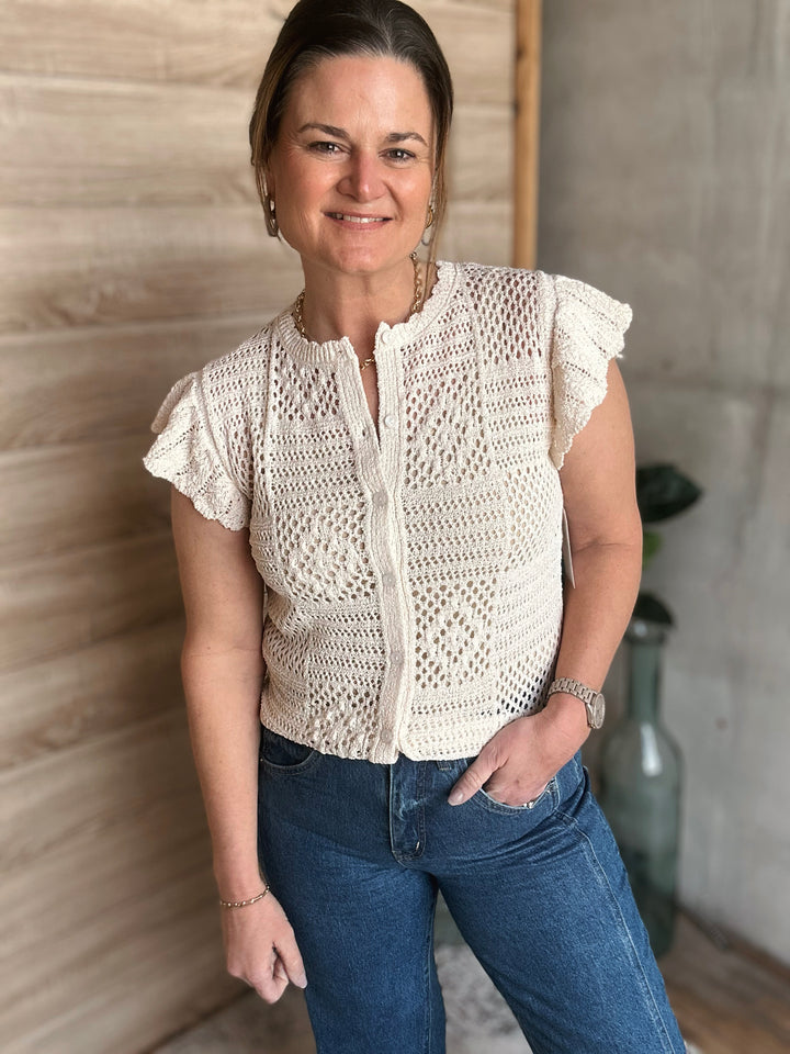 Woman wearing a white crochet top and blue jeans standing against a wooden wall.