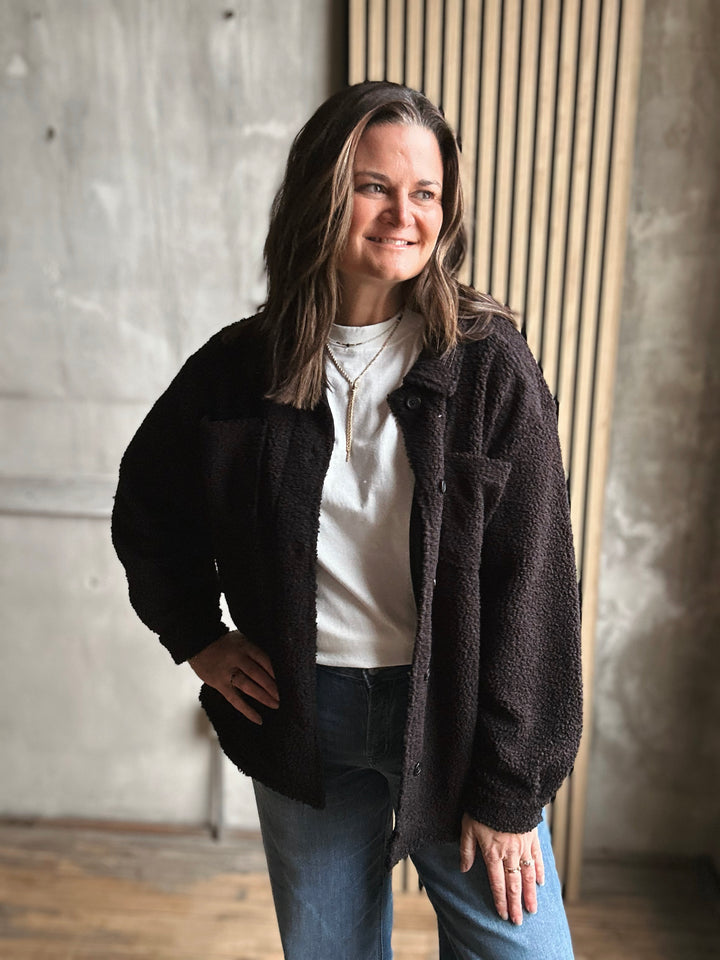 Woman wearing a black cardigan over a white shirt and blue jeans, standing against a neutral wall.