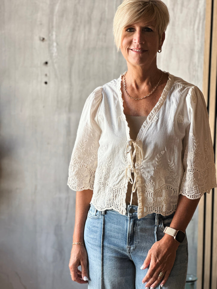 Woman wearing a white blouse with lace details and blue jeans against a neutral background