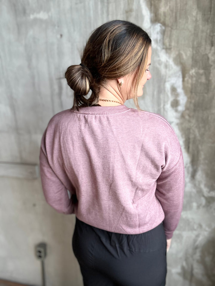 A woman viewed from behind wearing a dusty rose long-sleeve cropped sweatshirt and black pants, standing against a textured concrete wall.
