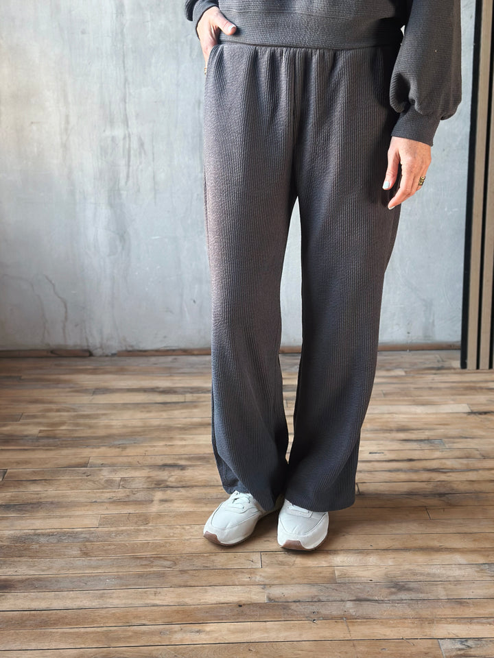 Person wearing dark gray pants and a matching top against a plain wall.
