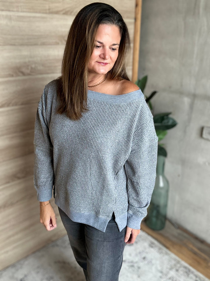 A woman wearing a light gray ribbed knit off-the-shoulder sweater and dark gray jeans, standing in an indoor setting with a wood-paneled wall and concrete wall in the background.