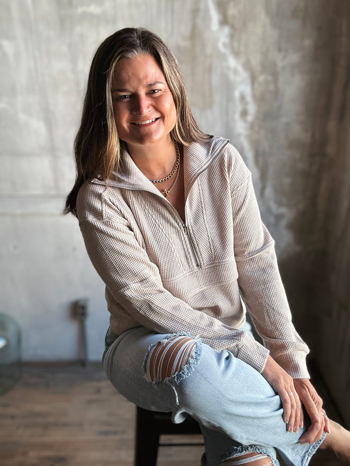 Woman wearing a beige hoodie and ripped jeans sitting against a textured wall.