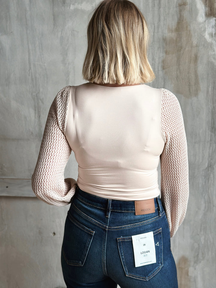 Person wearing a beige long-sleeve top and dark jeans with a visible brand tag, standing against a gray concrete wall.