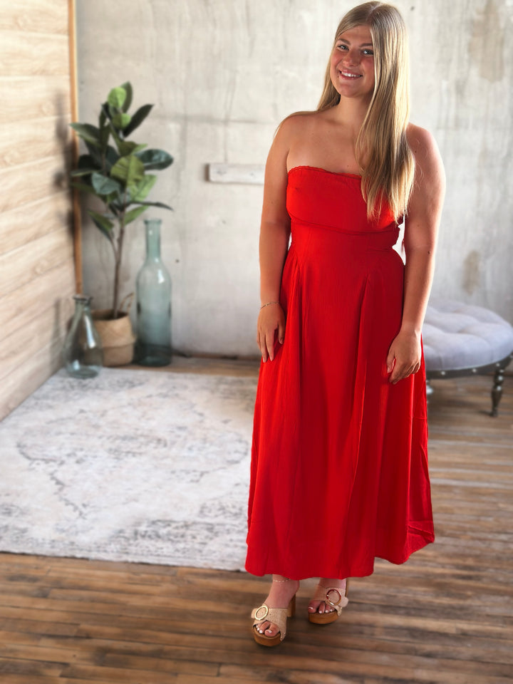 Woman wearing a red strapless dress standing in a room with wooden floor and white walls.