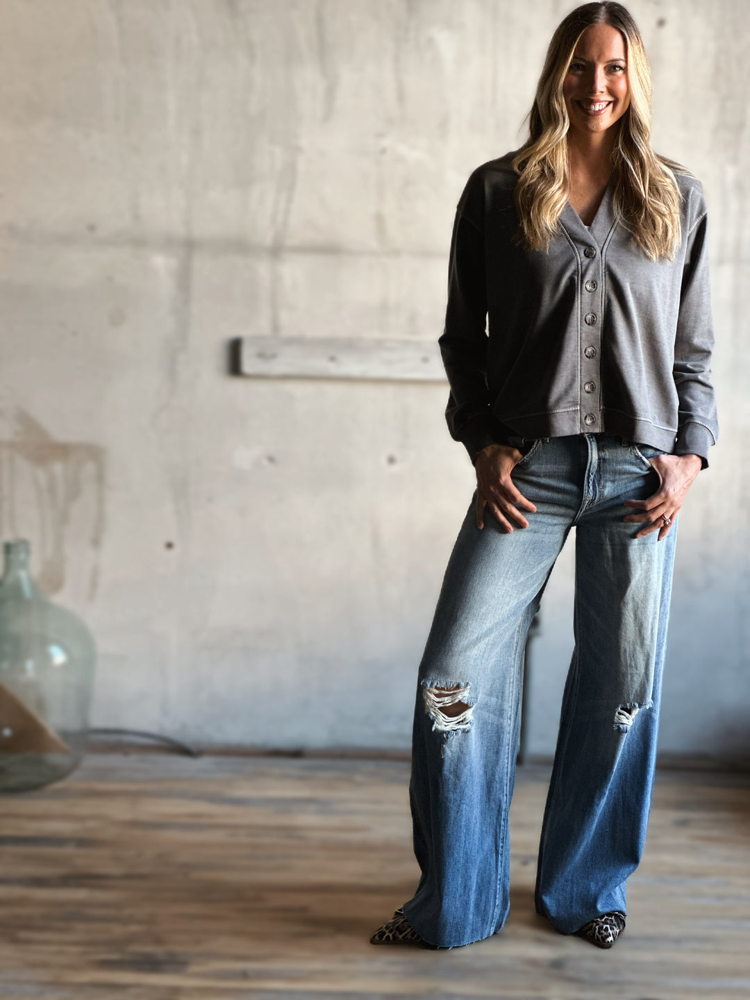 Woman wearing a gray button-up shirt, black jacket, and blue jeans in a room with a concrete wall.