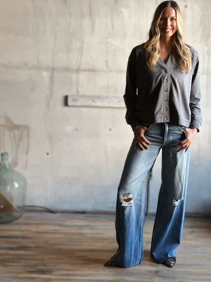 Woman wearing a gray button-up shirt, black jacket, and blue jeans in a room with a concrete wall.