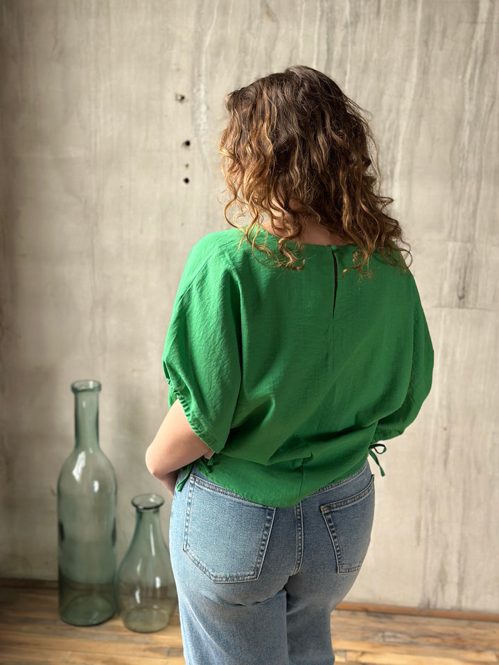 Person wearing a green blouse and blue jeans standing in front of a textured wall with two glass bottles.