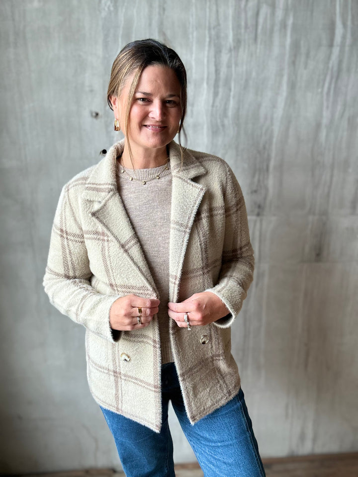 A woman wearing a beige and brown plaid wool blazer over a beige knit sweater and blue jeans, standing against a textured gray concrete wall.