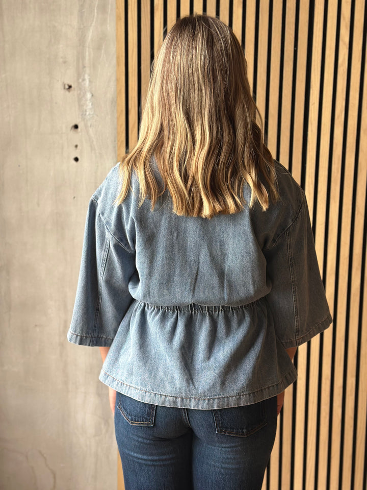 A woman viewed from behind wearing a light blue denim peplum blouse with three-quarter sleeves and dark blue jeans, standing in front of a slatted wood wall.
