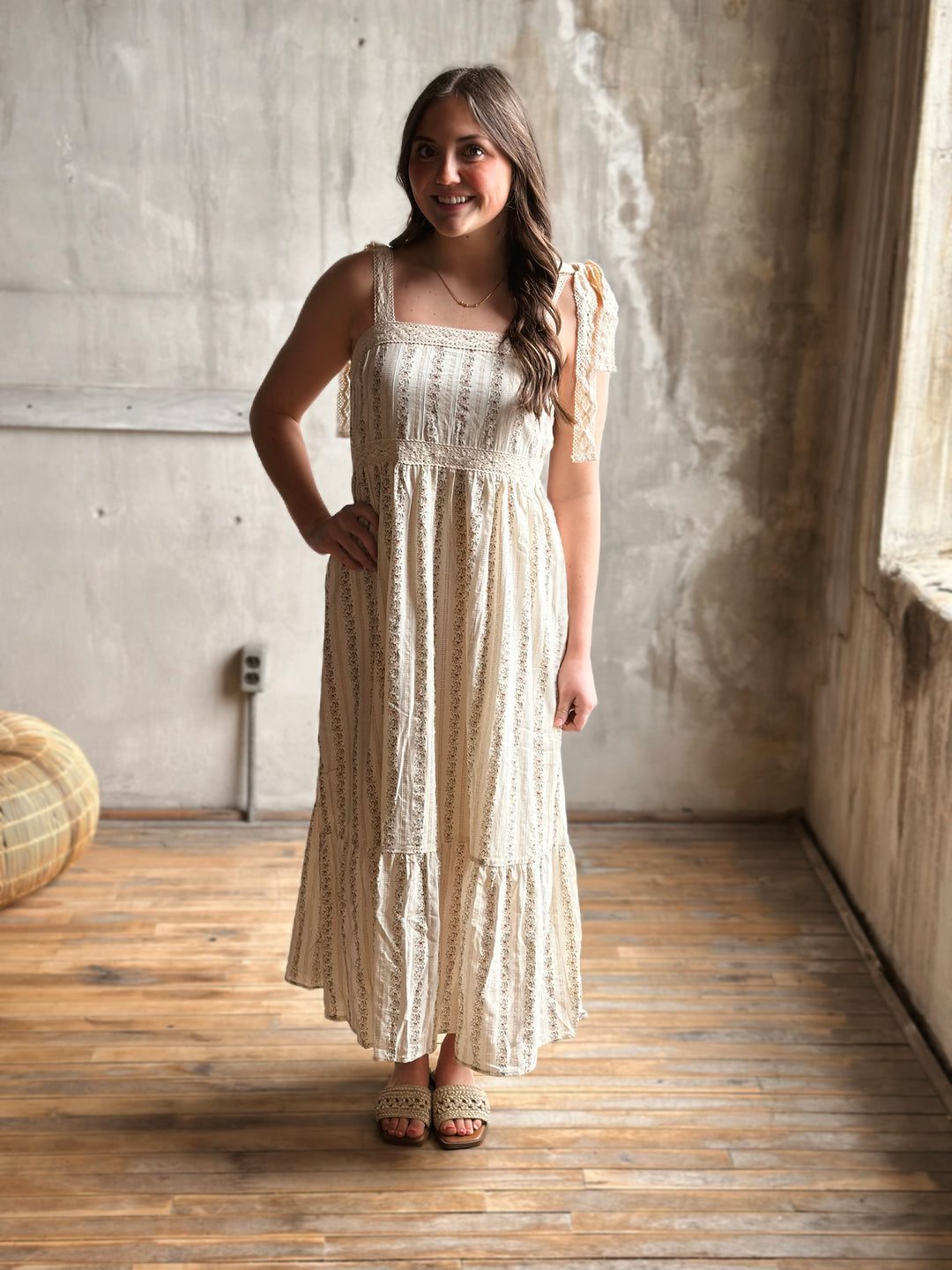 Woman in a long, light-colored dress with floral designs standing in a room with wooden flooring and a concrete wall.